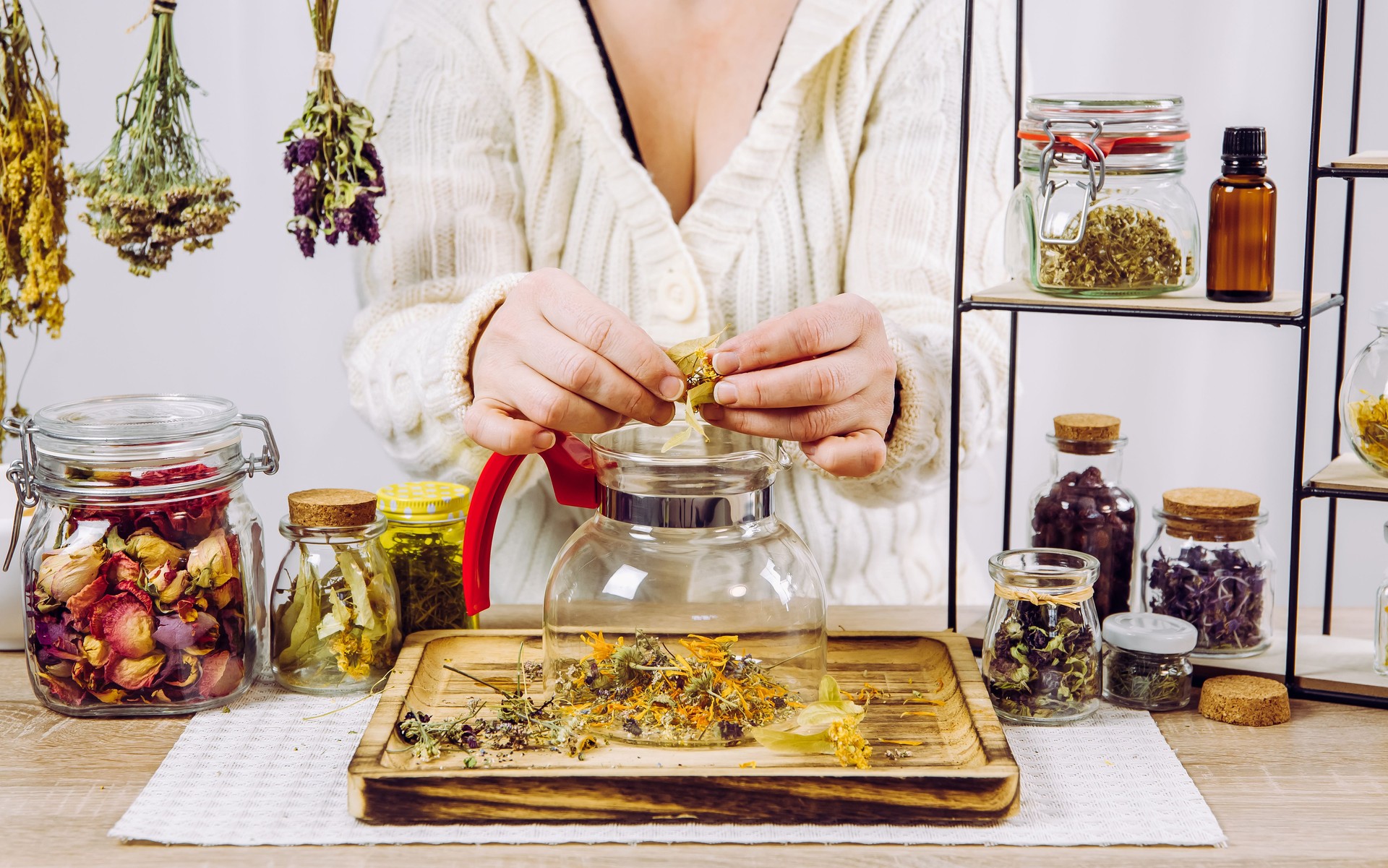 Close up view of woman herbalist mixing various dried herbs for traditional medicinal tea. Dried herbs in glass jars on background.