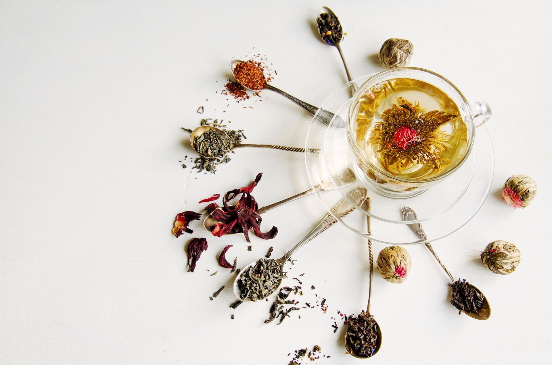 Blooming or flowering tea in a glass cup and silver spoons with various kinds of tea on white background